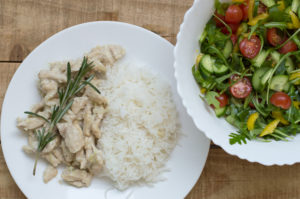White rice with chicken slices in white sauce and with fresh rosemary and big salad bowl with cucumber, arugula, cherry tomatoes and yellow capsicum. Balanced eating concept.