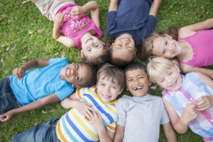 A multi-ethnic group of elementary age children are lying in the grass at the park. They have their heads together in the middle of a circle and are smiling and looking up at the camera.