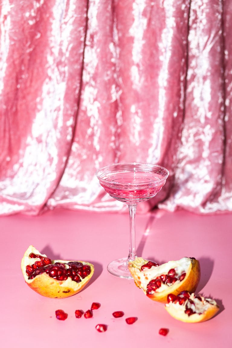 Elegant pomegranate cocktail in a coupe glass with pomegranate seeds on a pink velvet background.