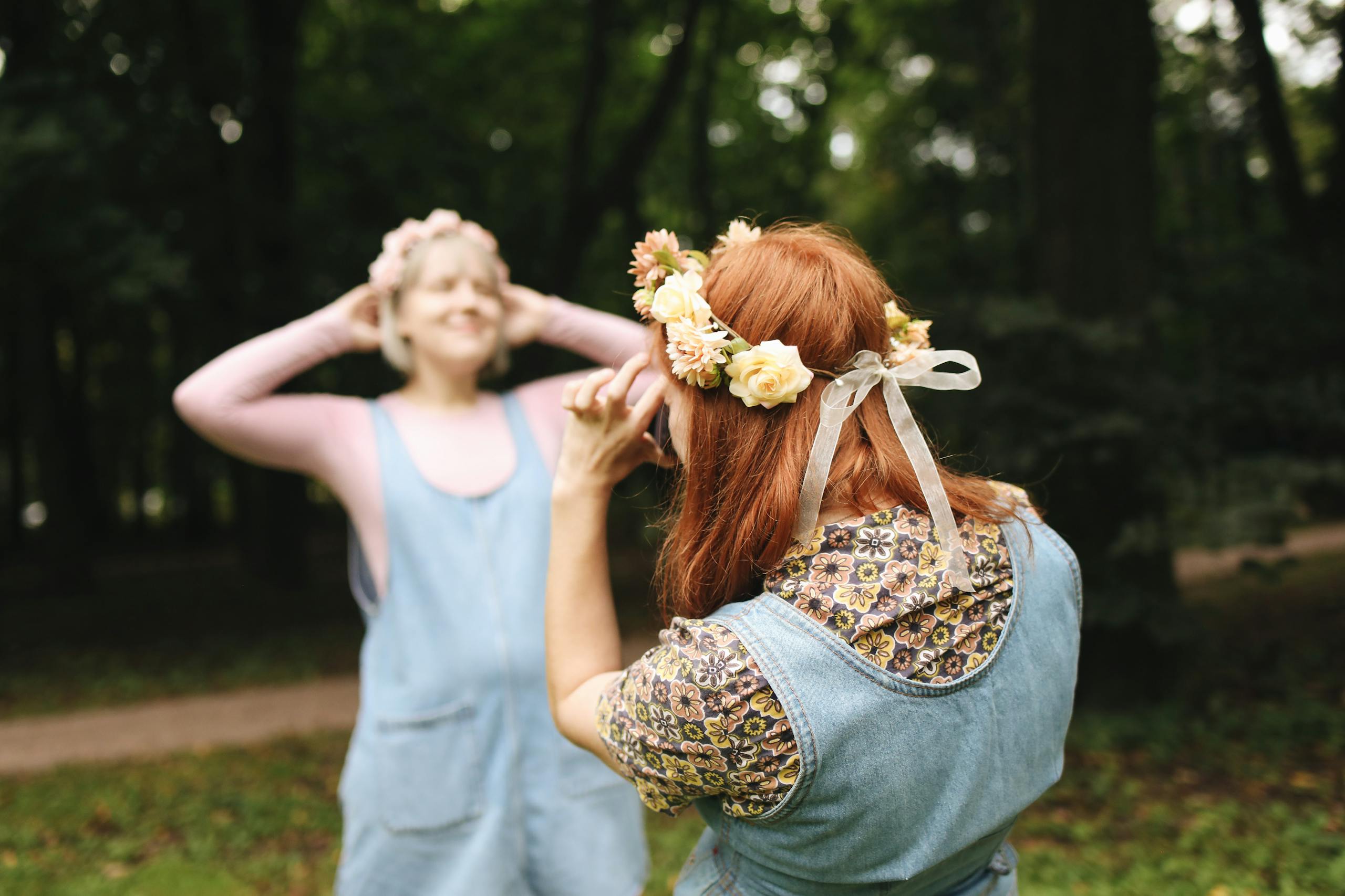 Two women in denim dresses and flower headbands posing playfully in a lush, green park.